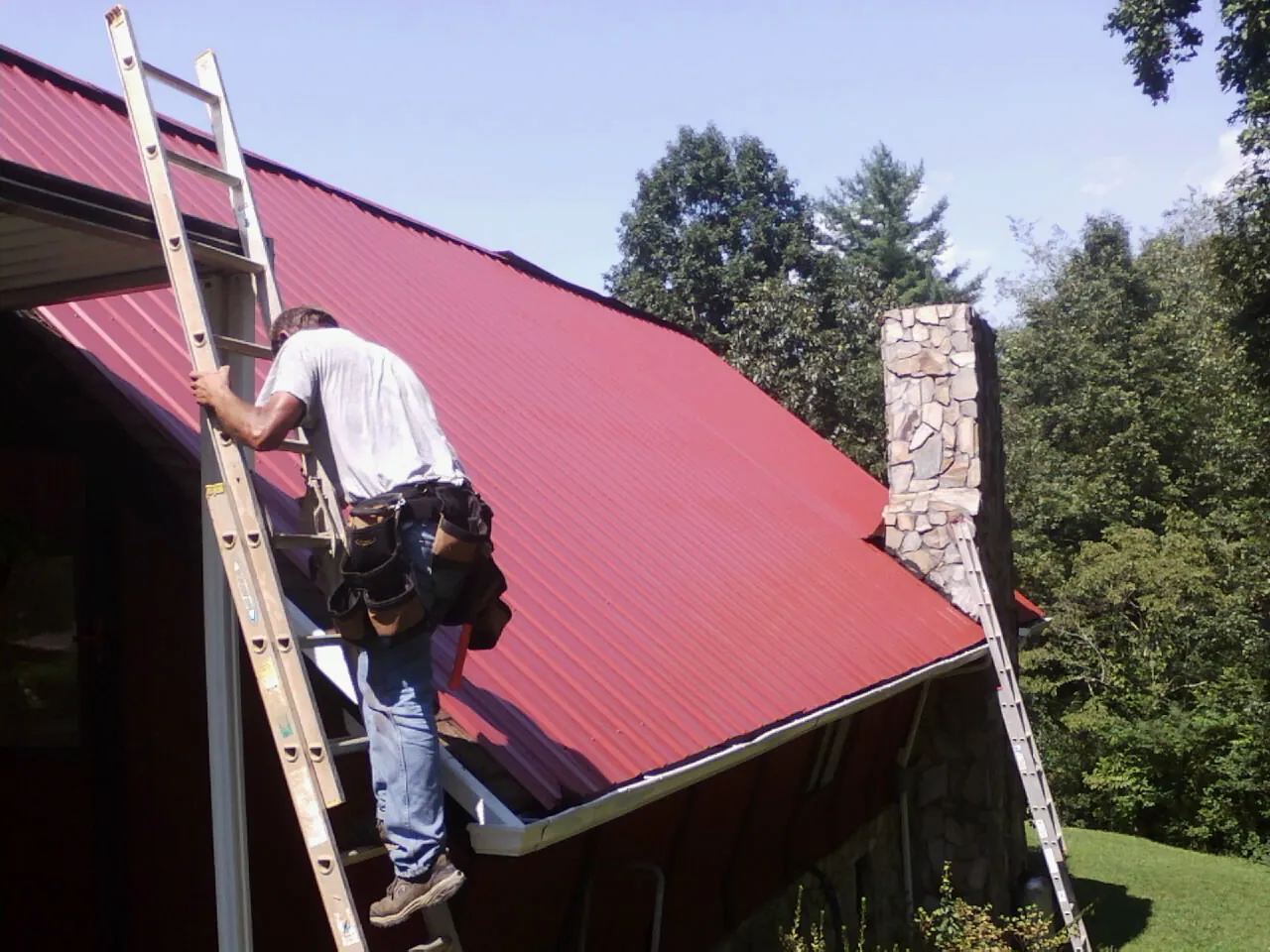 Roofer on a ladder beside a red metal roof and stone chimney — Soffit & Fascia Repair by Roofing Brothers in Barnegat Light